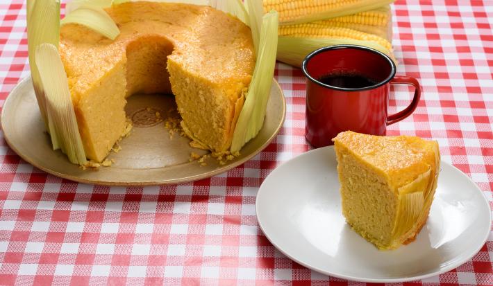 Foto de uma mesa com toalha vermelha quadriculada com dois pratos redondos, um com um bolo de fubá sem uma fatia, decorado com as folhas da espiga, e outro com uma fatia cortada em frente a uma xícara de café de louça vermelha