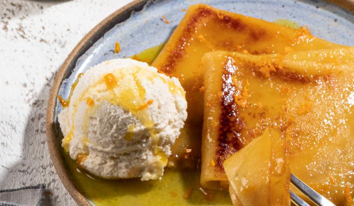 Foto de um prato em tom claro com a receita de Crepe Suzette, servido com calda amarela e uma bola de sorvete. O prato está em uma bancada clara, com. um pote de calda amarela e um guardanapo ao lado.