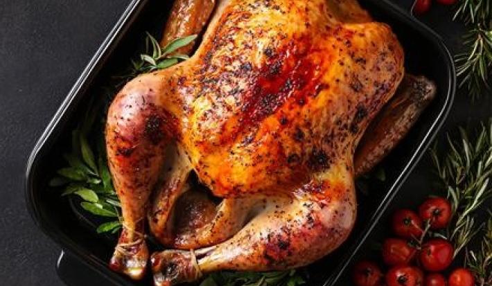 A imagem mostra um frango assado em uma travessa escura com ervas em sua volta