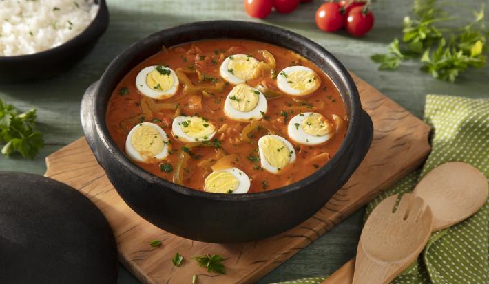 Fotografia em tons de verde em uma bancada de madeira verde, uma tábua de madeira, um paninho verde com bolinhas brancas, uma panela de barro grande ao centro com a moqueca de ovos dentro dela. Ao fundo, um recipiente com arroz, tomates e salada verde.
