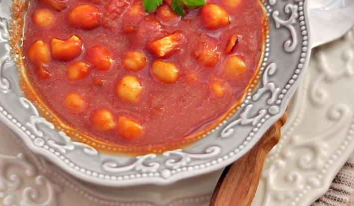 Fotografia vista de cima de uma sopa de tomates com nhoquettes em um prato fundo desenhado em tons de cinza, apoiado sobre um prato raso branco desenhado com uma colher apoiada. Os pratos estão sobre uma toalha de mesa marrom.