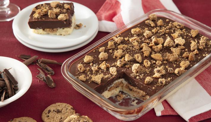 Fotografia de um pavê de chocolate com biscoito em uma travessa retangular transparente, com pedaços de biscoitos polvilhados por cima. Ao fundo uma toalha de mesa vermelha, com um prato de sobremesa branco servind um pedaço do pavê.