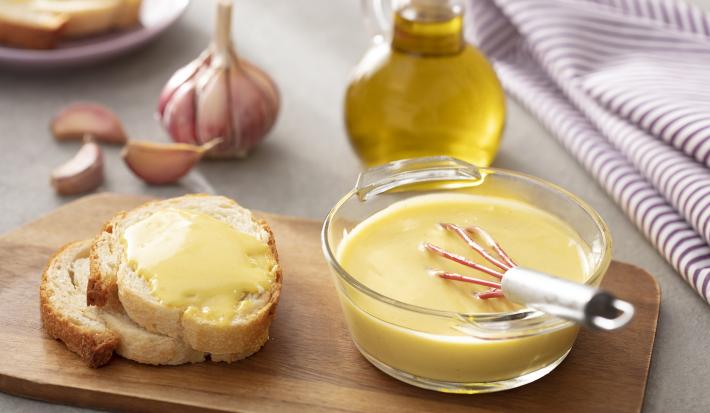 Foto em tons de amarelo da receita de aioli servida em um refratário de vidro redondo com um fuet mergulhado e fatias de pão ao lado com o o aioli, tudo sobre uma tábua de madeira. Ao fundo há um vidro de azeite e uma cabeça de alho