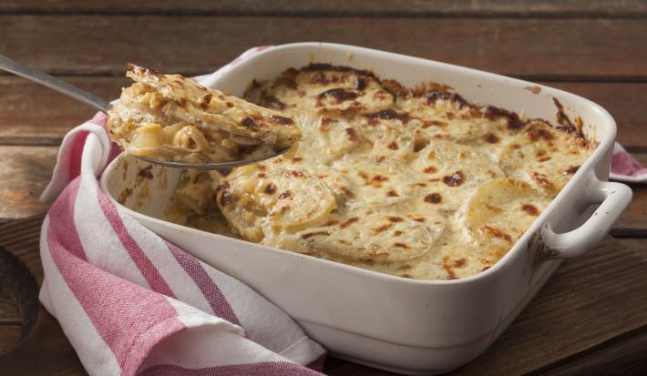 Fotografia em tons de marrom, branco e rosa de uma bancada marrom vista de frente. Contém um recipiente quadrado branco com alças e batatas e queijo gratinado, por cima uma colher com um pedaço da receita e ao lado um pano listrado nas cores rosa e branco