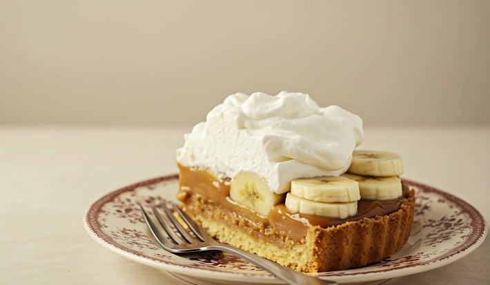 Fotografia em tons claros de uma fatia generosa de banoffee em um prato pequeno e desenhado sobre uma mesa clara. Ao lado da fatia e sobre o prato tem um garfo apoiado.