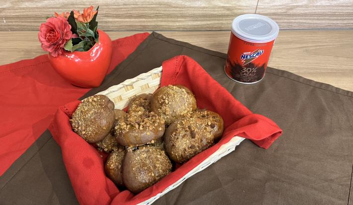 Fotografia de pãezinhos de nescau com castanhas dentro de uma cestinha branca quadrada com um pano vermelho. A cestinha está sobre um pano marrom de tom escuro, ao lado da latia de nescau extra cacau.