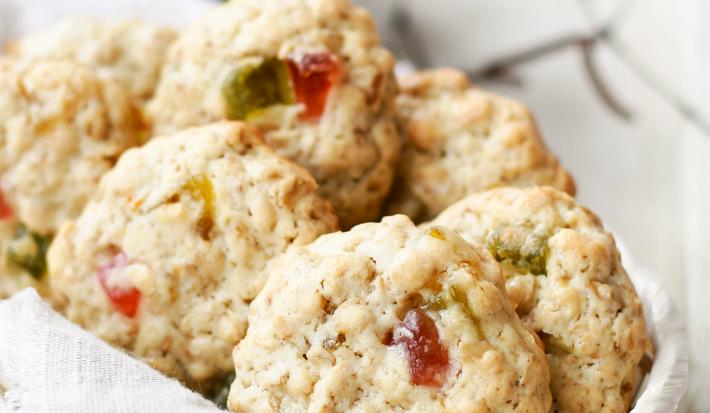Fotografia em tons claros de cookies de massa branca e neston e frutas que estão dentro de uma cestinha de madeira forrada com um pano branco. Ao fundo, um copo de alumínio branco com desenhos em azul e verde. Tudo está sobre uma mesa de madeira clara.
