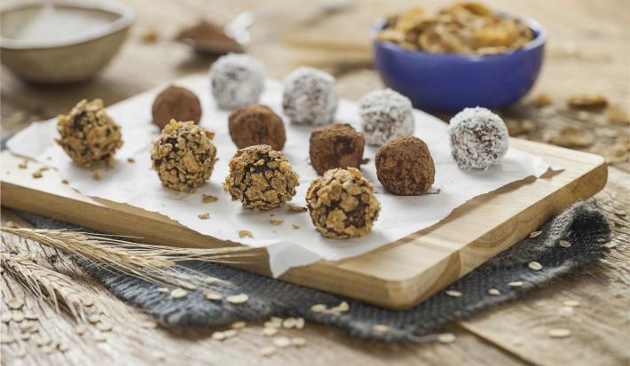 fotografia em tons de azul em uma bancada de madeira clara, uma tábua de madeira, um paninho azul escuro e vários docinhos parecidos com brigadeiros, mas feitos com Nesfit e sem açúcar. Ao fundo, potinhos com Nesfit Cereais.