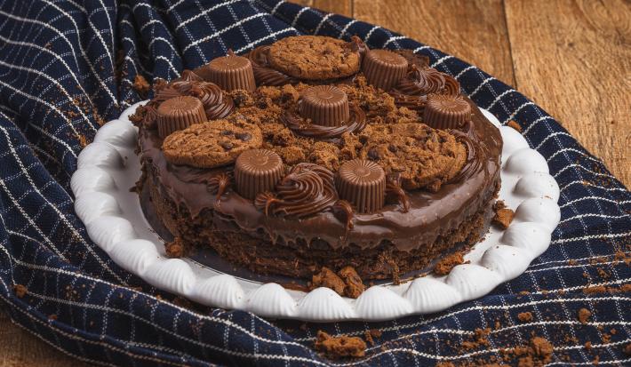 Foto de uma bancada de madeira. Sobre ela há uma toalha azul escuro com listras brancas e um prato branco com a receita da Torta Creme Alpino decora com bombons e cookies.