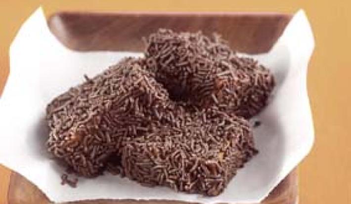 Fotografia em tons de marrom em uma bancada de madeira, uma bandeja de madeira escura, com um papel toalha embaixo e três marias moles de chocolate, com chocolate granulado por cima.