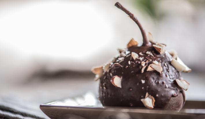 Fotografia de uma pera coberta com chocolate à base de Neston e lascas de nozes sobre um prato de vidro de cor clara.