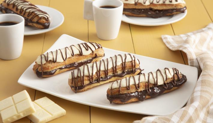 Fotografia em tons de amarelo em uma bancada de madeira com um pano xadrez brege, um recipiente retangular branco ao centro com três bombas de chocolate com cobertura de chocolate branco. Ao fundo, duas xícaras com café e dois pratinhos com duas bombas.