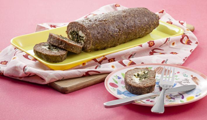Fotografia em tons de rosa em uma mesa rosa com um pano rosa desenhado e um recipiente retangular amarelo com o bolo de carne com espinafre em cima dele. Ao lado, um prato colorido com uma fatia do bolo e um garfo e uma faca apoiados sobre ele.