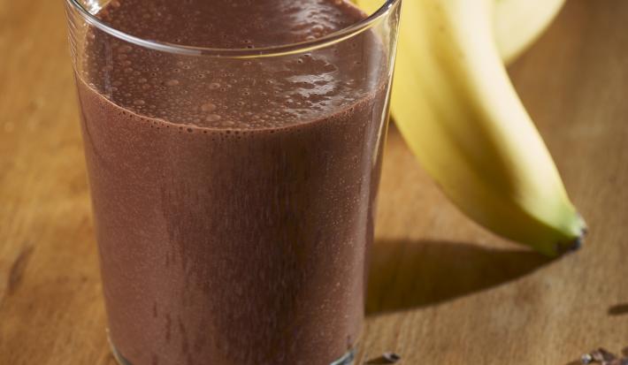 Fotografia de um copo de vidro com uma vitamina de banana e chocolate Nescau. Ao fundo, um cacho de bananas, e na frente do copo, raspas de chocolate sobre uma mesa de madeira.