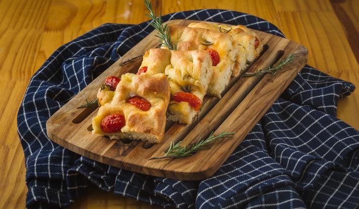 Foto aproximada de uma bancada com um tecido azul quadriculado sobre e nele uma tábua de madeira com pedaços de Focaccia, decorada com tomates-cereja e ramos de alecrim