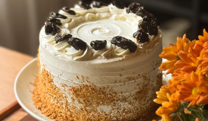 Fotografia de um bolo inteiro de doce de leite com ameixa e coco, decorado com chantilly e ameixas sobre um prato grande e fundo branco.
