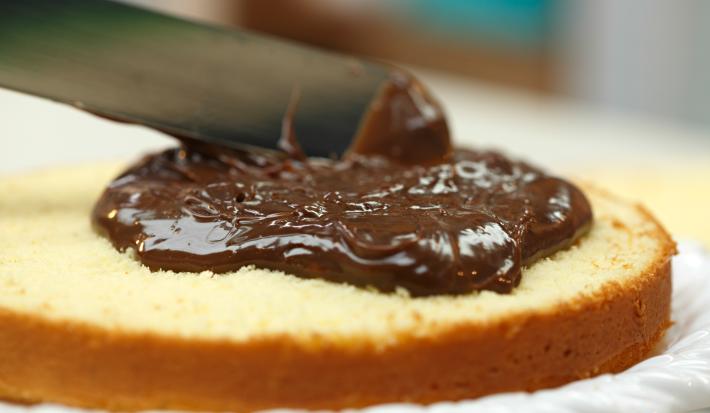 Imagem de um bolo redondo branco cortado com um recheio de brigadeiro sendo espalhada pela espátula.