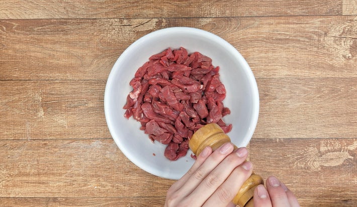 Mãos temperando carne bovina com um moedor de pimenta-do-reino preta moída em uma tigela branca sobre uma bancada de madeira.