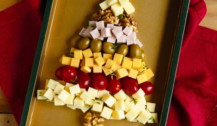 Fotografia em tons de vermelho em uma bancada de madeira escura, um pano vermelho, uma bandeja com vários frios (muçarela, presunto, queijo prato, tomatinhos e nozes) empilhados formando uma árvore de natal.