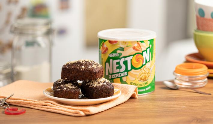 Foto de uma bancada de madeira. Sobre ela há uma tecido laranja claro e nele um prato pequeno com três brownies de cacau. Ao fundo há uma lata de Neston 3 Cereais e alguns utensílios de cozinha