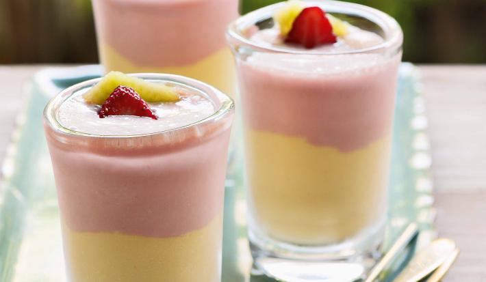 Fotografia em tons de rosa e amarelo em uma bancada de madeira, um recipiente retangular com três taças de vidro com o creme de frutas com o sorvete de creme. Morango e manga para decorar as taças.