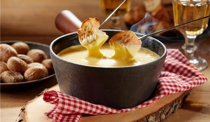 Fotografia mostra um creme brilhante com textura aveludada, servido em aparelho de fondue de cerâmica, acompanhado de cubos de pão dourados mergulhado