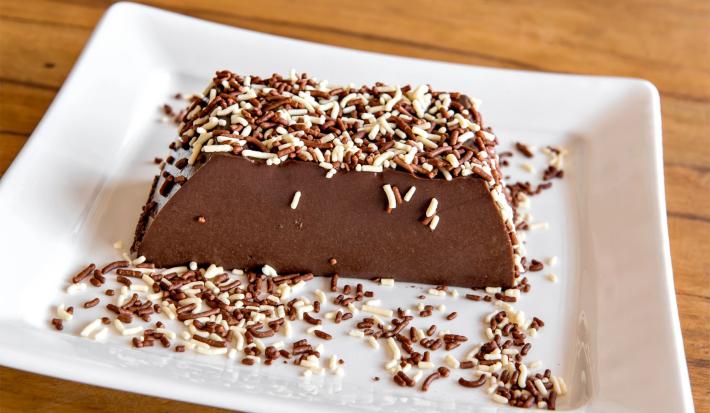 Imagem de um pedaço da sobremesa de chocolate coberta com chocolate granulado branco e preto sobre um prato.