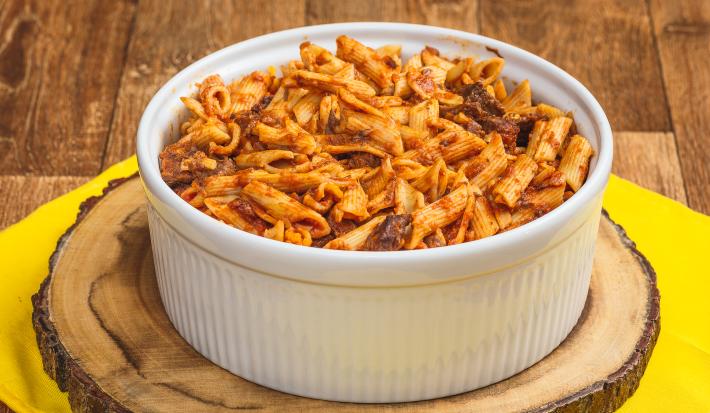 Foto de uma bancada de madeira e bem ao centro há uma toalha amarela, uma tábua de madeira e sobre ela um recipiente redondo e branco. Dentro dele está a receita de macarrão tipo penne com carne desfiada.