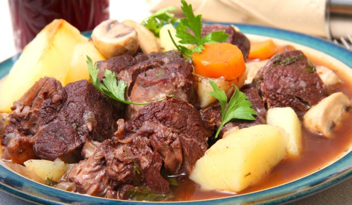 Fotografia com um prato branco e bordas na cor azul. Dentro, um cozido de carne, batata, cenoura e champignon, com folhas de salsinha por cima.