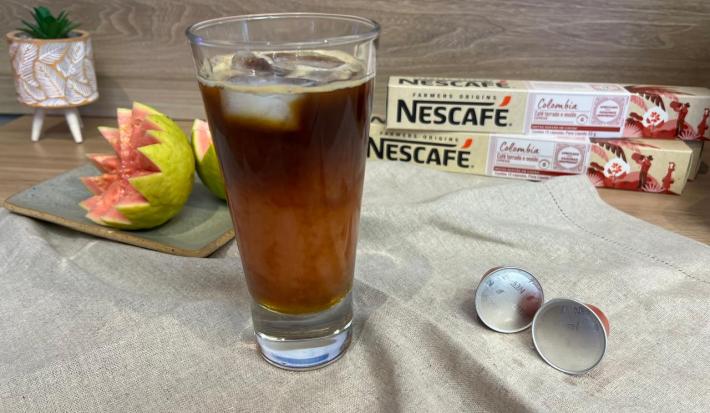 Fotografia em tons de rosa e verde com um copo alto ao centro. Dentro do copo existe uma mistura de café com suco de goiaba e cubos de gelo. Ao fundo existe uma goiaba cortada e duas caixas de cápsulas de NESCAFÉ