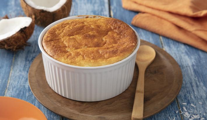 Foto da Receita de Suflê de Coco. Observa-se um recipiente redondo com o suflê sobre uma tábua de madeira redonda. Ao lado do potinho, uma colherinha também de madeira. Ao fundo, um coco aberto e um guardanapo laranja ambientam a foto.