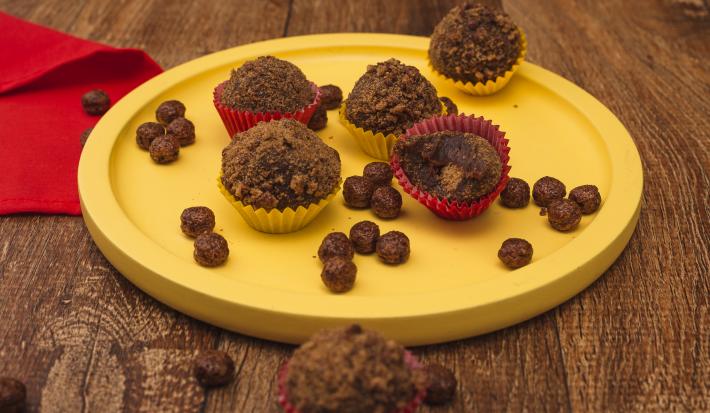 Fotografia em tons de amarelo em uma bancada de madeira escura, um pano vermelho, um prato amarelo redondo raso com brigadeiros de Nescau e recheados com Nescau Cereal.