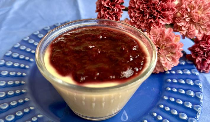 Fotografia em tons de azul e rosa com um pote transparente ao centro. Dentro do pote existe uma receita de flan de mamão coberto com geleia de frutas.