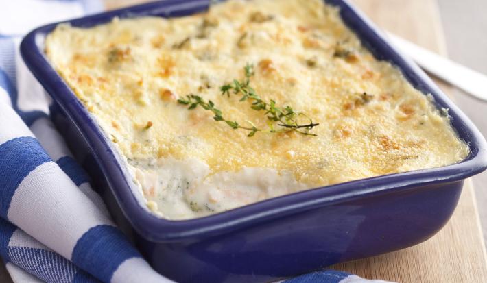 Fotografia em tons de azul em uma bancada de madeira clara, um pano branco com listras azuis, uma tábua de madeira, um recipiente quadrado azul com os legumes gratinados dentro dele.