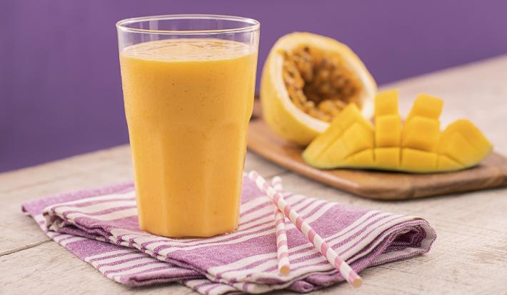 Foto em tons de roxo e amarelo da receita de smoothie de manga e maracujá servida em um copo de vidro largo sobre um pano lilás listrado com dois canudos ao lado. Ao fundo uma tábua de madeira com maracujá e manga cortados ao meio.