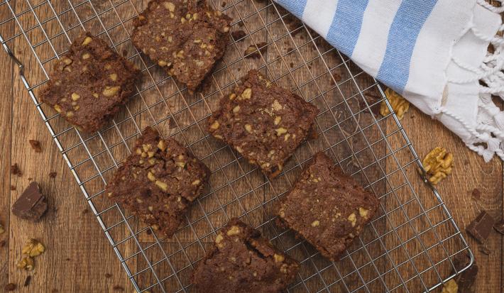 Fotografia em tons de azul em uma bancada de madeira escura, um suporte de grelha prata com alguns brownies de chocolate com nozes em cima dele e ao lado um paninho branco com listras azuis.