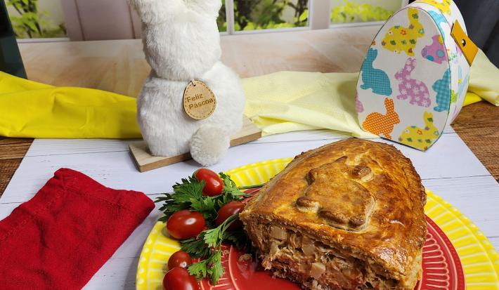Foto da receita de Ovo de Colher Torta de Frango, servido cortado em um prato vermelho, sobre outro prato amarelo, em uma bancada branca decorada com itens de páscoa, como um coelho branco e uma caixinha em formato de ovo de páscoa