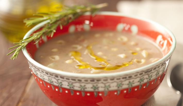 Fotografia em tons de vermelho em uma mesa de madeira com um recipiente redondo e fundo vermelho com a sopa de feijão dentro. Ao lado, um guardanapo de pano bege com uma colher apoiada nele e ao fundo, um potinho de azeite.