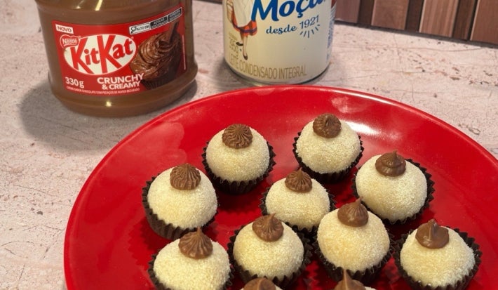 Docinhos cobertos com coco ralado em prato vermelho, decorados com creme de KitKat e Leite Moça ao fundo.