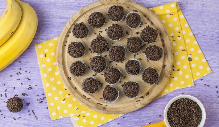Fotografia em tons de marrom, amarelo e lilás, com bancada lilás vista de cima, ao centro peça de madeira com brigadeiros sobre guardanapo amarelo com bolinhas brancas, ao lado potinho com granulado, um brigadeiro e e duas bananas.