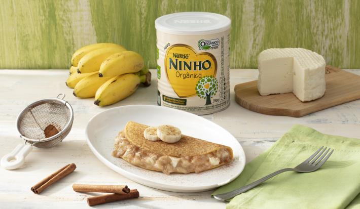 Fotografia em tons de branco e verde de uma bancada branca com um prato branco redondo com uma panqueca. Ao lado um paninho verde com um garfo, paus de canela, uma peneira, um cacho de banana, um queijo branco e uma lata de Ninho Orgânico.