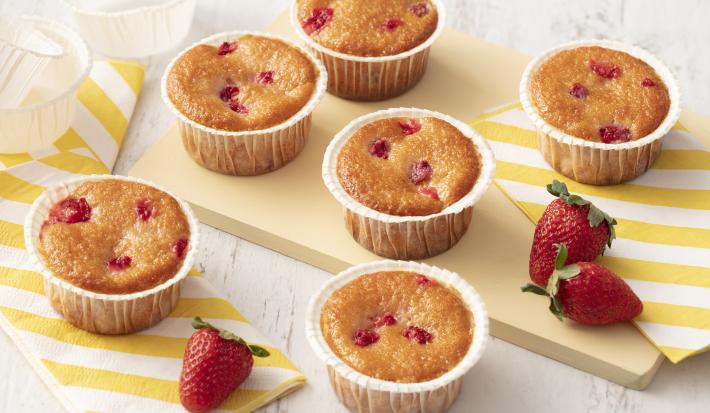 Fotografia em tons de branco e amarelo de uma bancada de madeira branca com uma tábua de madeira, sobre ela um paninho listrado branco e amarelo, cupcakes e morangos. Ao lado outro paninho listrado amarelo e branco com morangos e dois cupcakes.