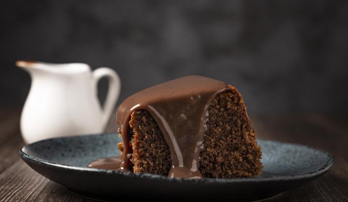 fotografia em tons de preto e marrom tirada de uma fatia de bolo de chocolate com calda por cima, e ao fundo um jarra branca