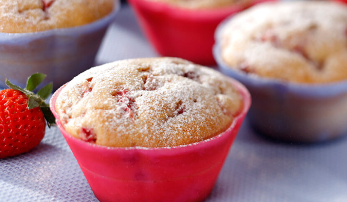 Fotografia em tons de vermelho e lilás em uma bancada com uma toalha emborrachada lilás, com forminhas pequenas vermelhas e lilás com os muffins de fubá com morango dentro delas.