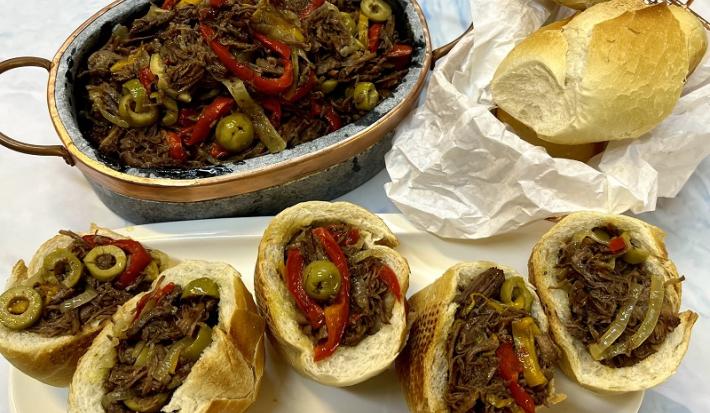 Foto da receita de carne louca servida em uma panela de pedra circular com 5 lanches de carne louca feitos a frente com pão francês e a receita dentro. Ao lado ainda há um saco de pães franceses