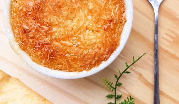Foto da receita de Cocada de Forno. Observa-se um recipiente redondo branco com a cocada dentro assada e bem douradinha, sobre uma tábua de madeira