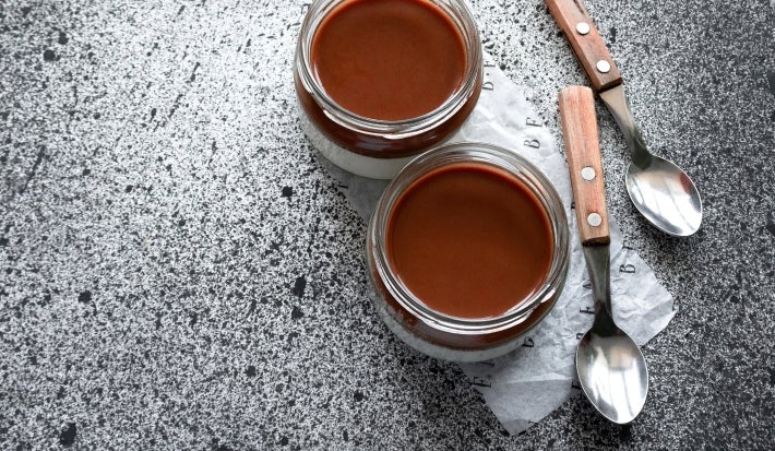 Fotografia vista de cima de dois recipientes de vidro, decorativos, com a sobremesa, finalizada com uma camada grossa de ganache. Ao lado dos recipientes tem duas colheres de sobremesa com o cabo de madeira.
