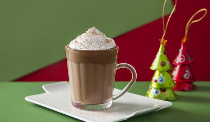 Fotografia em tons de marrom em uma bancada de madeira de cor verde. Ao centro, um bandeja branca contendo a bebida. Ao fundo, há 3 kikat sabor Natal.