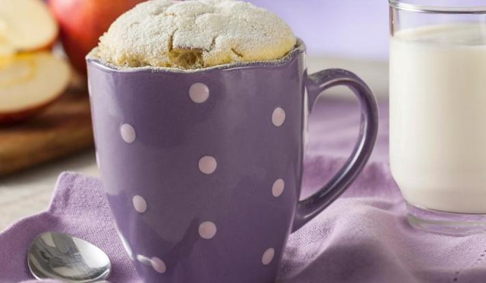 Fotografia em tons de roxo em uma bancada de madeira e um paninho roxo. Ao centro, uma caneca roxa de bolinhas brancas com o bolinho de caneca de leite Ninho em Pó. Ao fundo, maçãs cortadas ao meio e um copo de vidro com leite.
