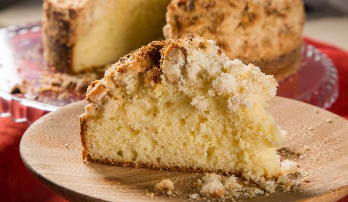 Fotografia de uma receita de cuca alemã tradicional com uma fatia grande cortada sobre um prato raso de madeira. No fundo tem a receita quase inteira sobre um apoio de vidro raso.